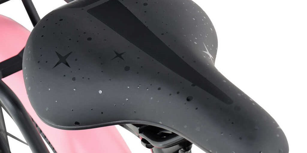 Vaaleanpunainen lasten 16" polkupyörä, jossa on matala jalannostokorkeus, etukori, takateline, seisontatuki, lokasuoja ja ketjusuoja. Lähikuva satulasta.